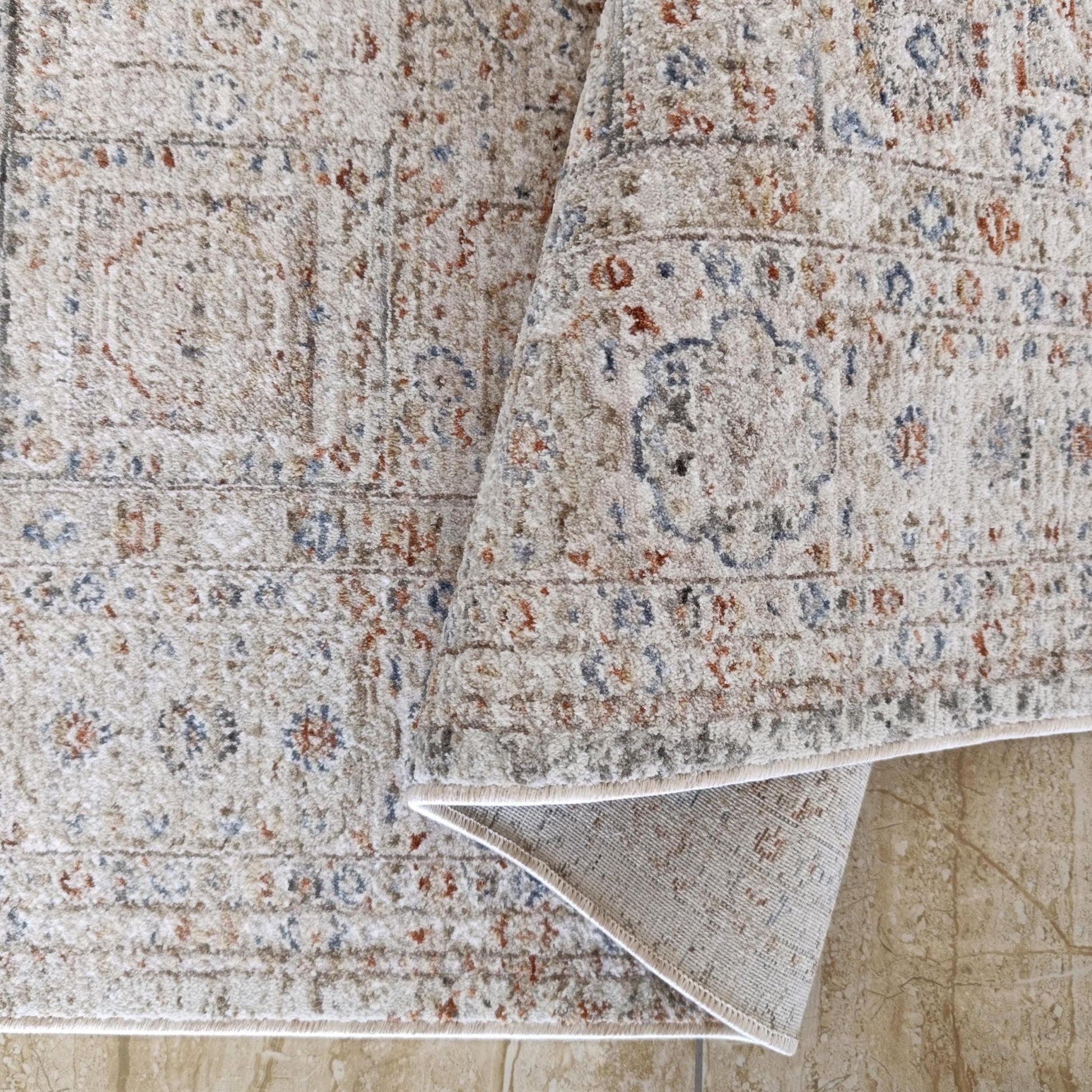 Tapis à poils clairs avec des motifs traditionnels bleus et rouille, partiellement roulé sur un sol en carrelage marbré beige