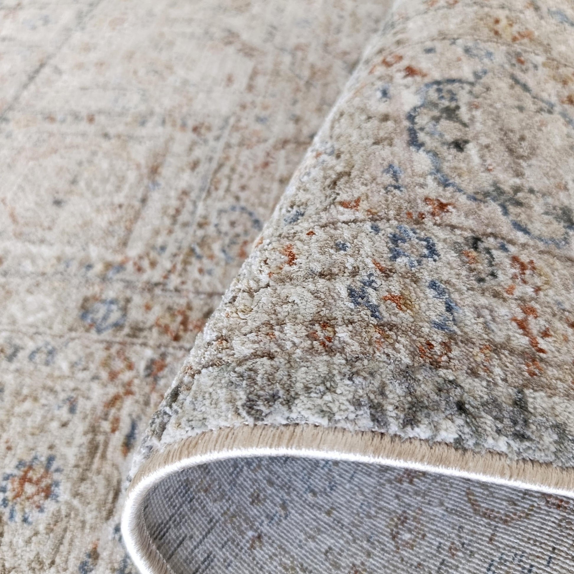 Tapis à poil court couleur sable aux motifs ornés gris-bleu et rouille, partiellement roulé, aspect vieilli