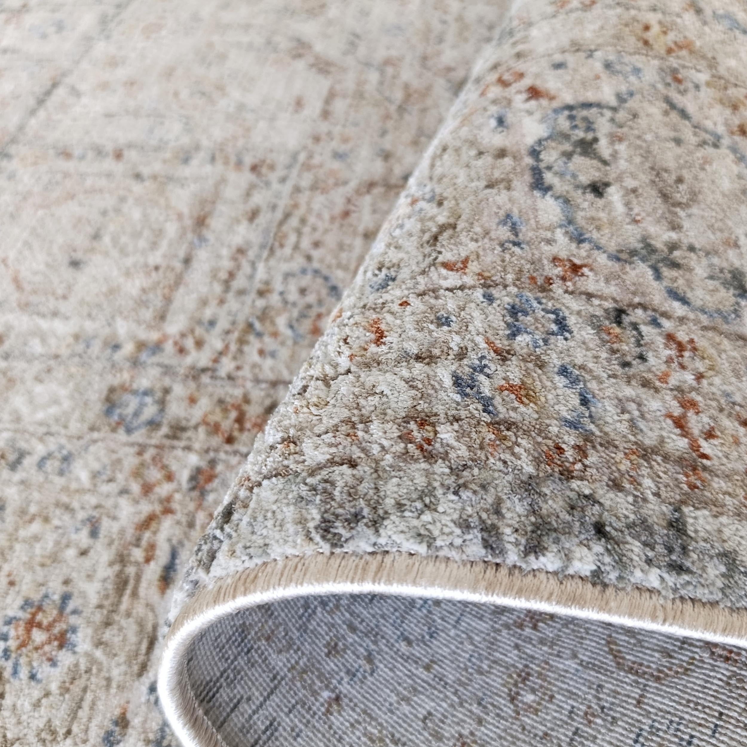 Tapis à poil court couleur sable aux motifs ornés gris-bleu et rouille, partiellement roulé, aspect vieilli