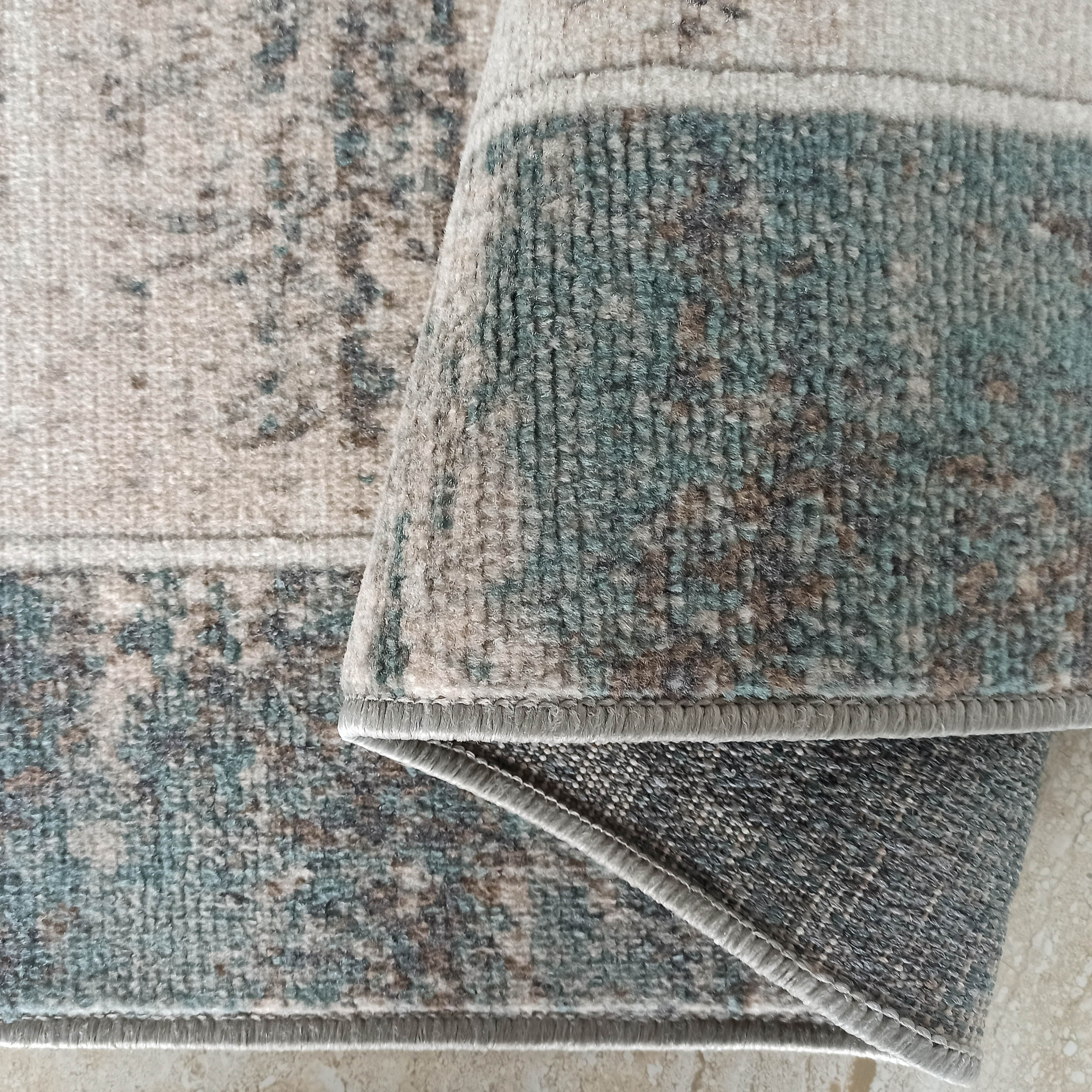 Gros plan d'un tapis partiellement roulé, aux motifs abstraits beige, turquoise et gris, avec son envers texturé