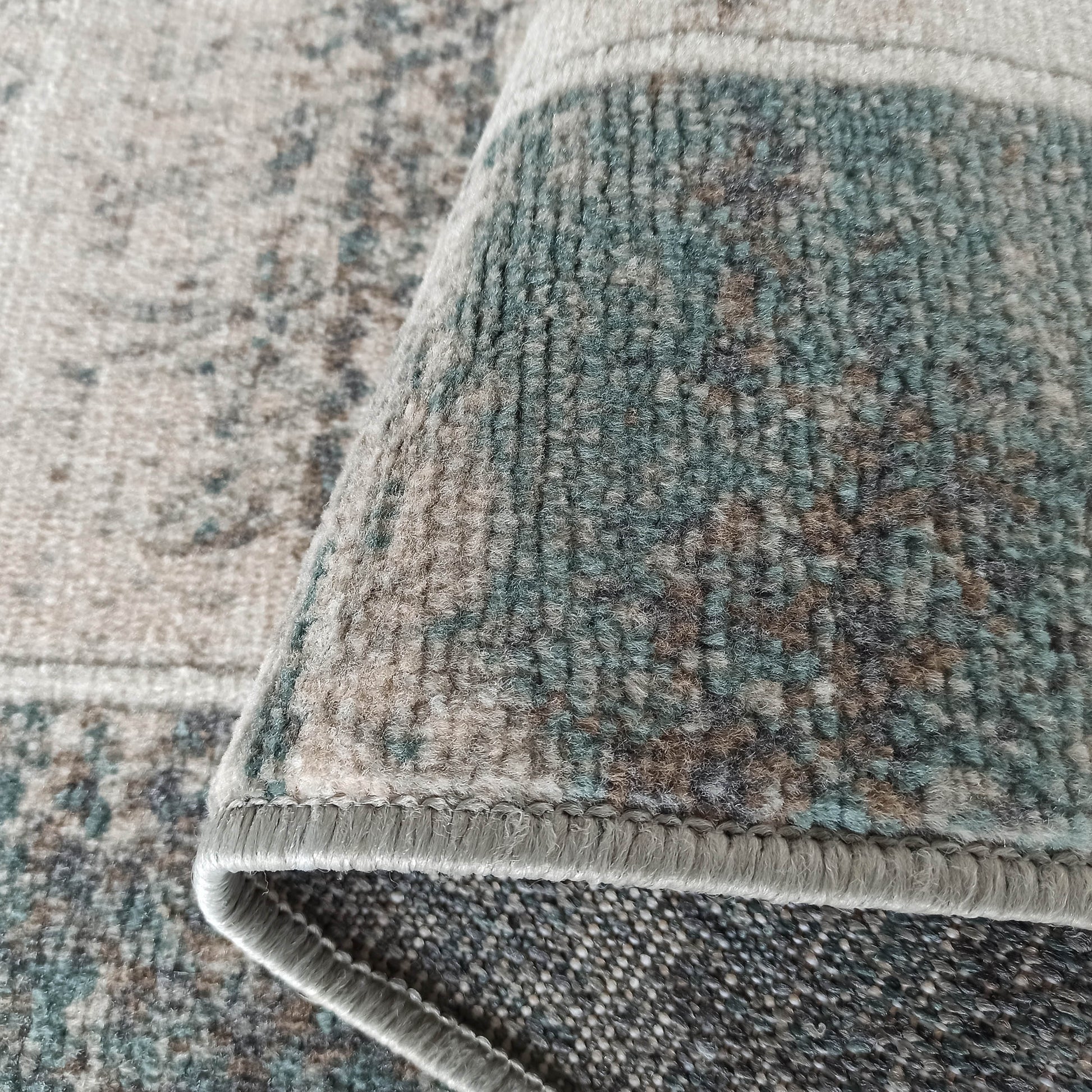 Gros plan sur un tapis à la texture douce et aux motifs abstraits beige, bleu-vert et marron, avec un bord replié