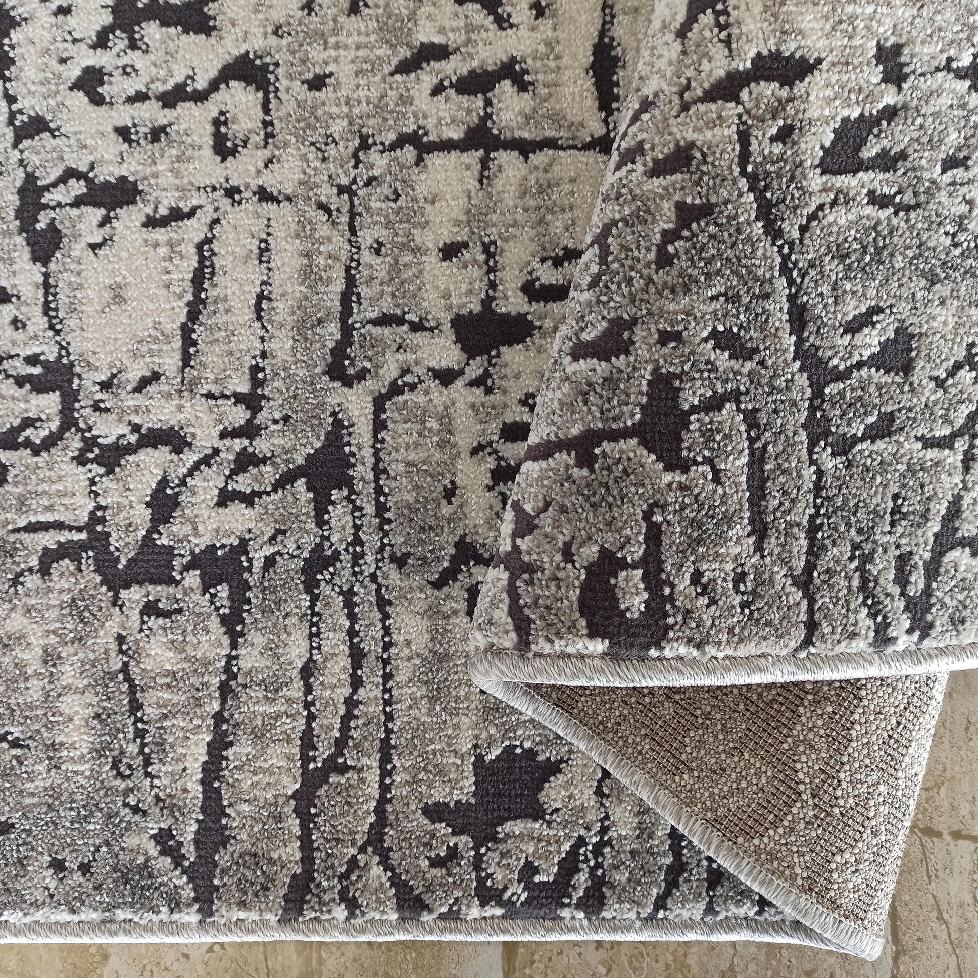 Tapis moelleux à motifs abstraits gris et anthracite, avec un coin replié montrant son envers texturé sur carrelage