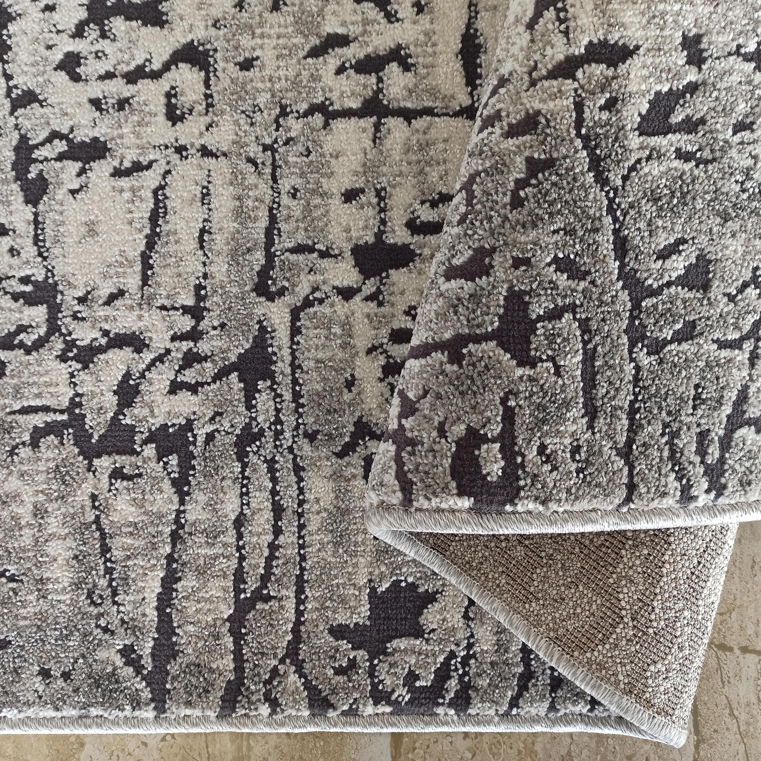 Tapis moelleux à motifs abstraits gris et anthracite, avec un coin replié montrant son envers texturé sur carrelage