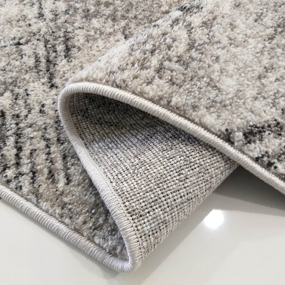 Gros plan sur un tapis gris clair à motifs anthracite, dont le côté en poil long est plié, révélant le revers tissé et le bord gansé