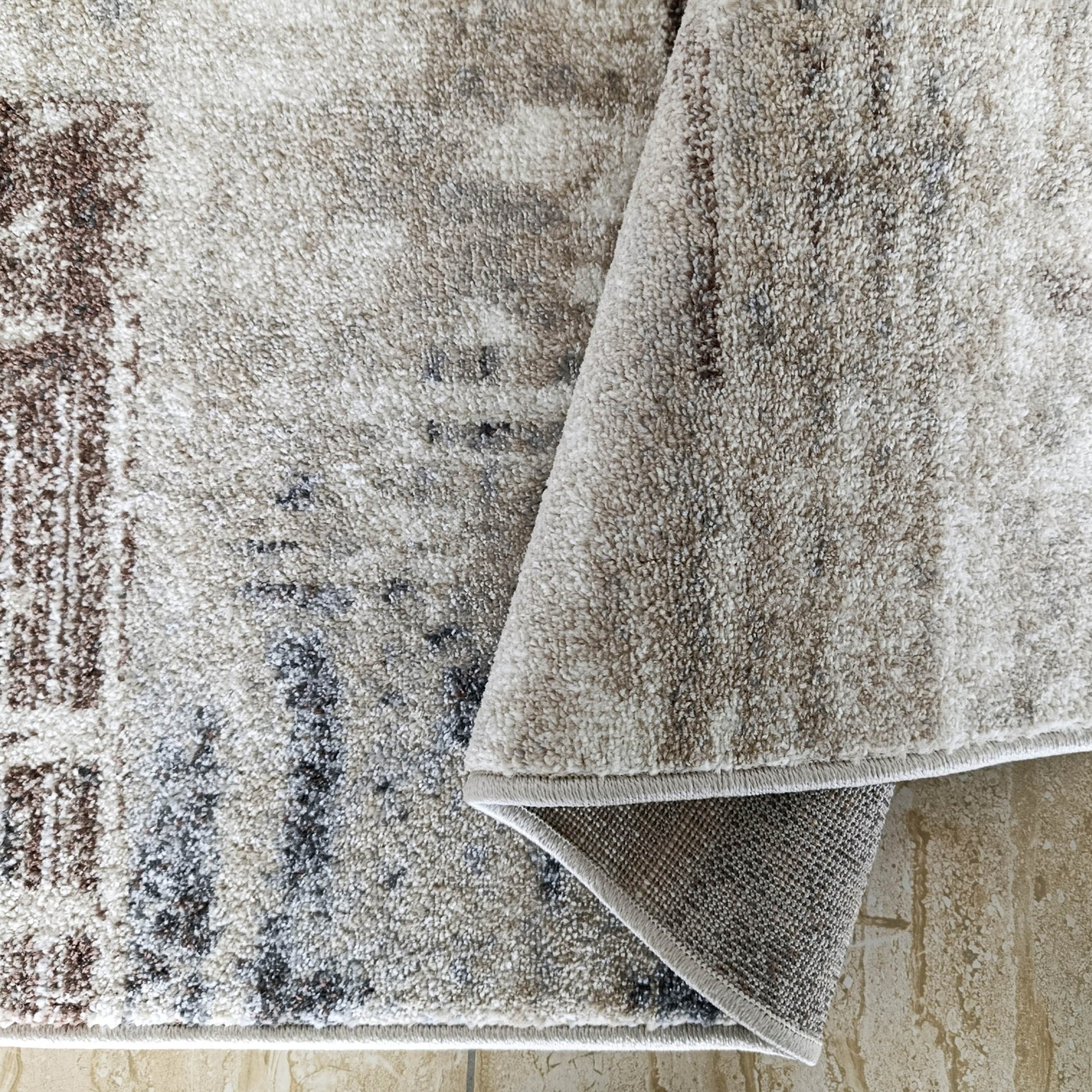 Tapis moelleux beige clair à motifs abstraits marron et gris foncé, partiellement enroulé sur un sol carrelé texturé