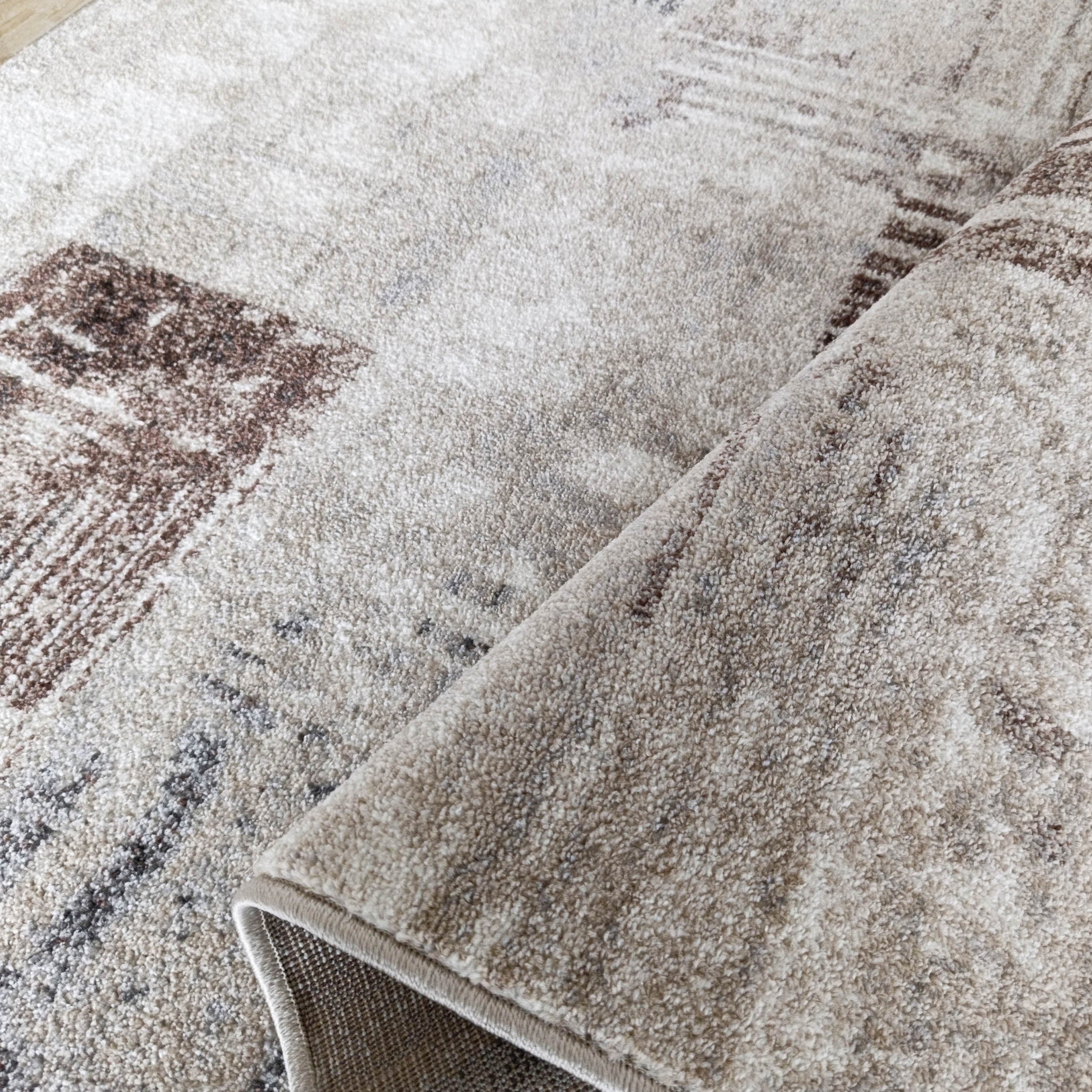 Tapis doux à poils courts beige clair, orné de motifs abstraits marron et gris, avec une partie de la bordure roulée