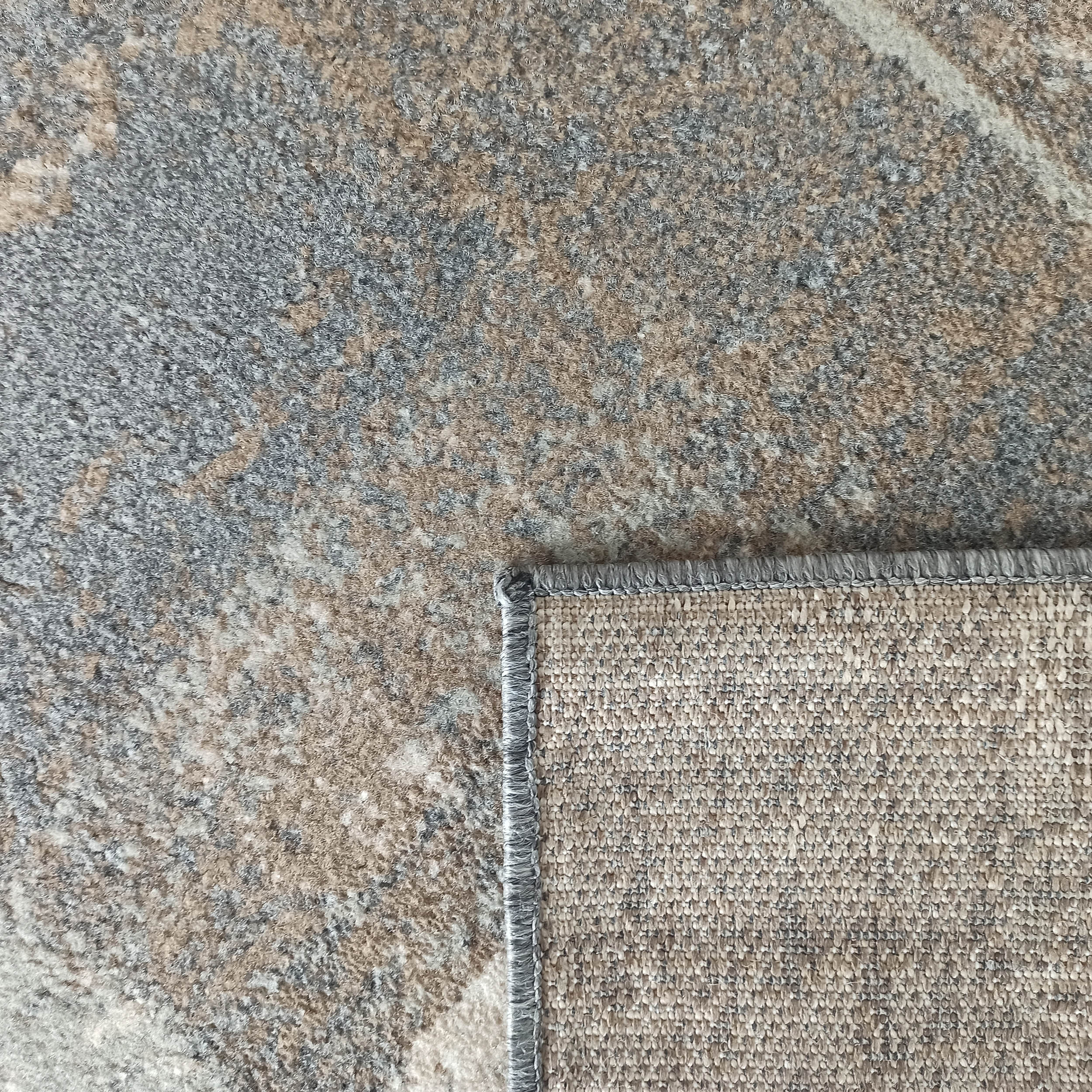 Tapis texturé aux teintes grises, beiges et blanches, montrant son envers tissé et sa bordure surpiquée