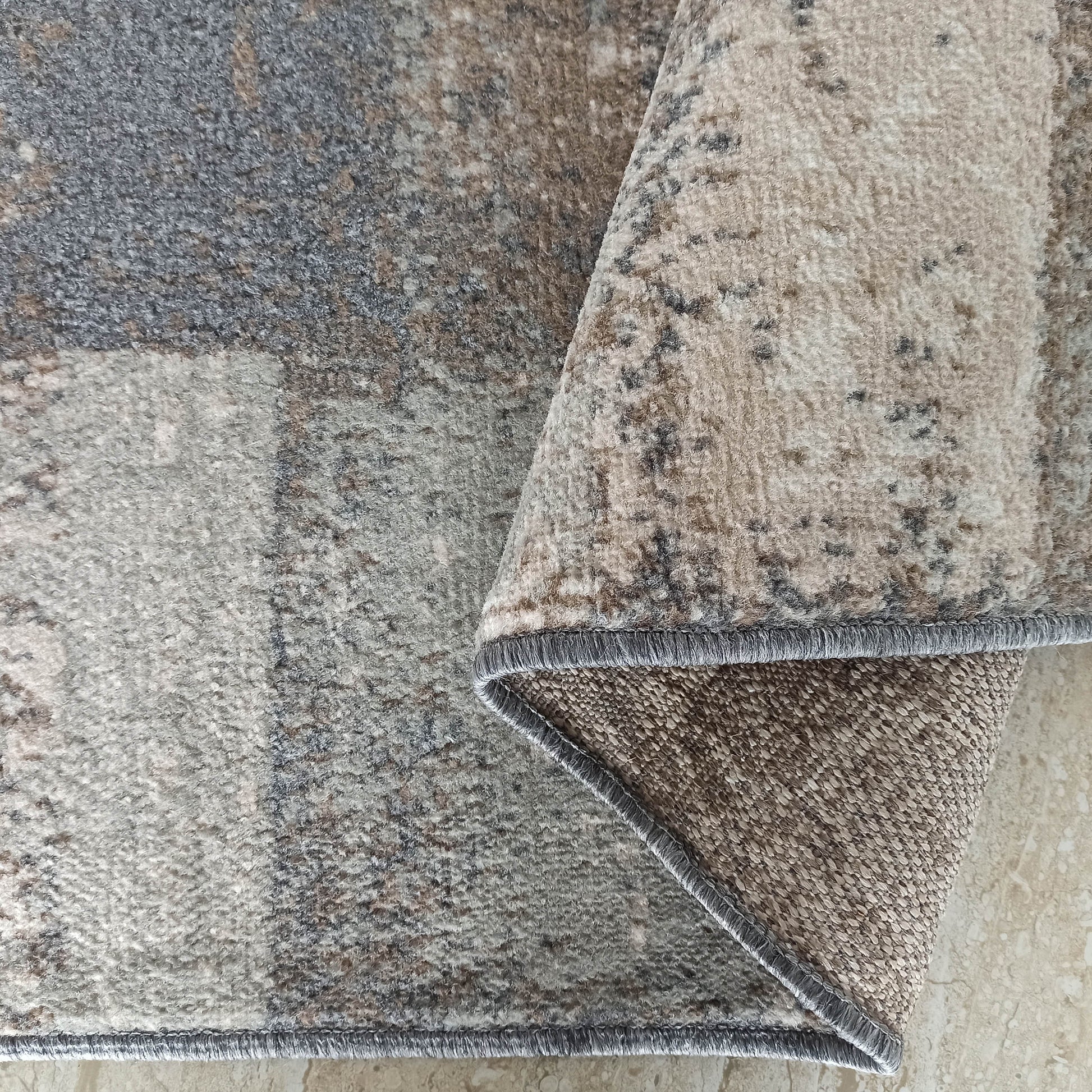 Tapis à la texture douce et aux motifs abstraits gris, beiges et bruns. Un coin est replié, révélant son envers et la bordure surjetée