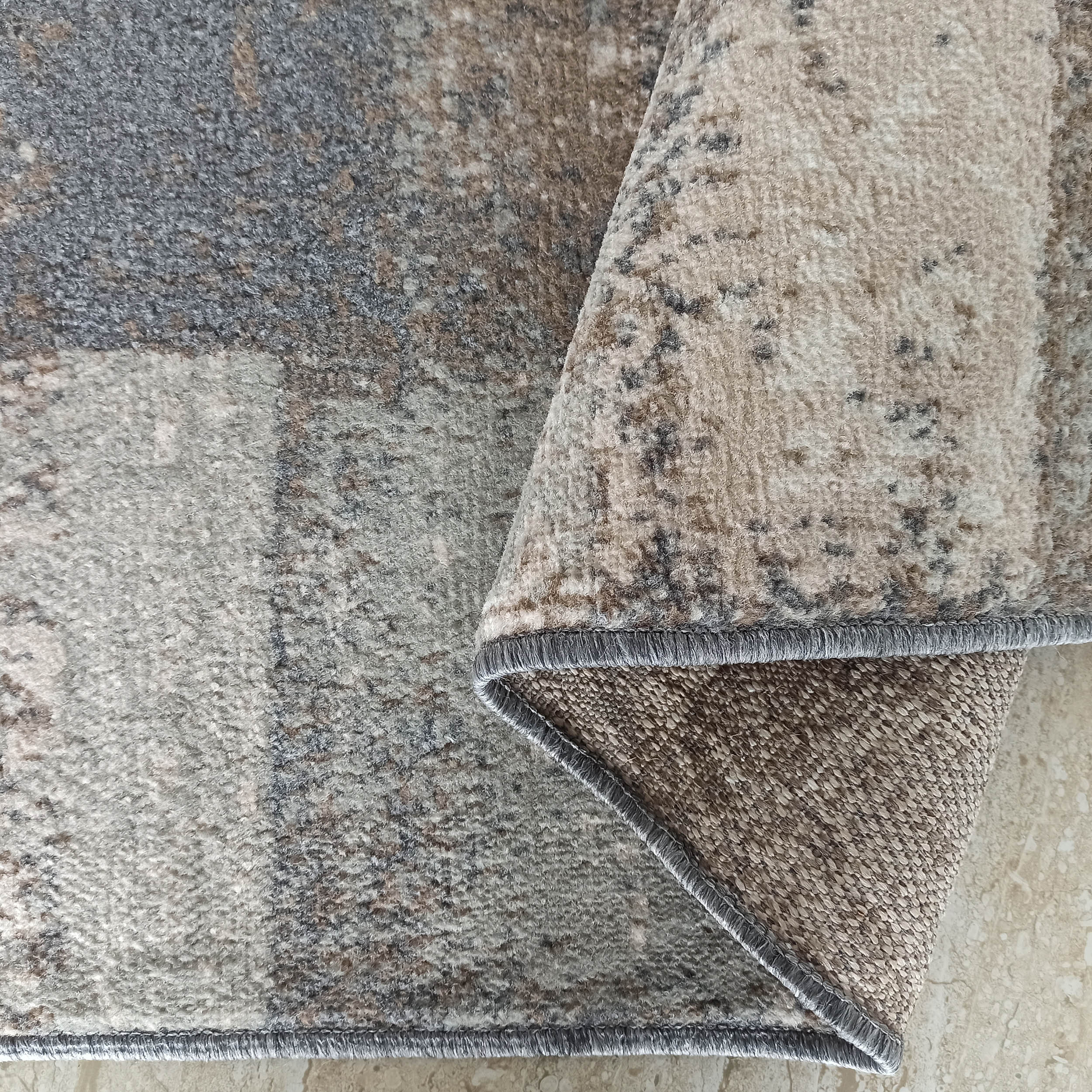 Tapis à la texture douce et aux motifs abstraits gris, beiges et bruns. Un coin est replié, révélant son envers et la bordure surjetée