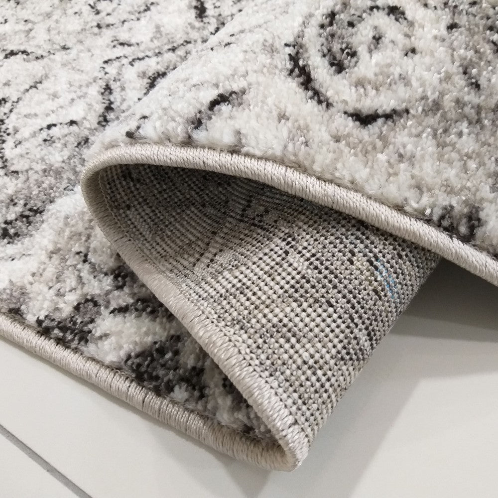 Gros plan sur un tapis à motifs abstraits blancs, gris et noirs, montrant sa texture duveteuse et son revers tissé gris avec une bordure cousue claire