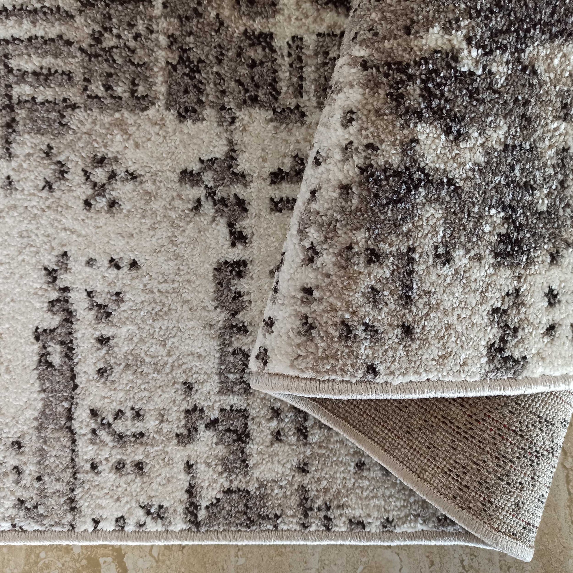 Tapis moelleux aux motifs abstraits gris foncé et beige clair, avec le dos tissé visible sur le côté roulé