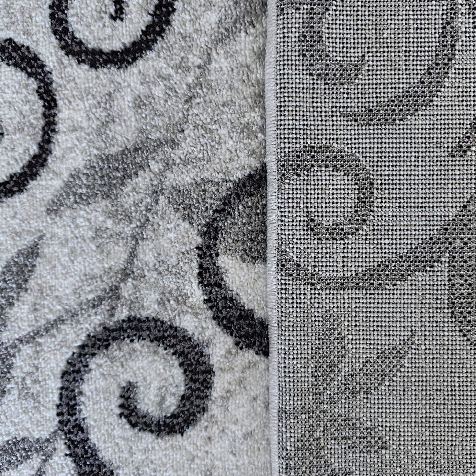 Tapis moelleux aux motifs de volutes grises et noires sur fond blanc-gris, avec sa sous-couche tissée à droite