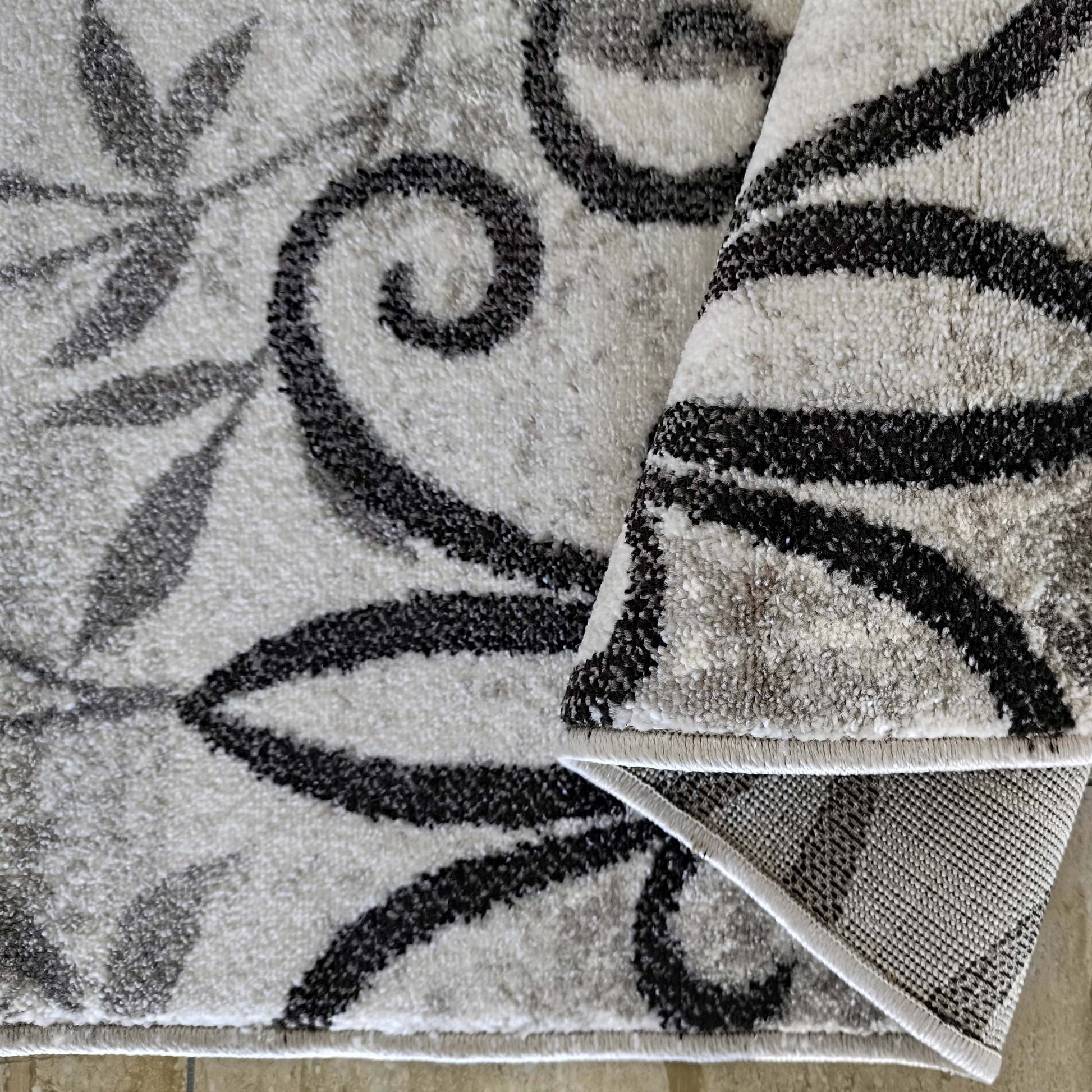 Tapis à poil court aux motifs de feuilles et volutes noirs et gris sur fond blanc et gris clair, roulé pour montrer son envers textile