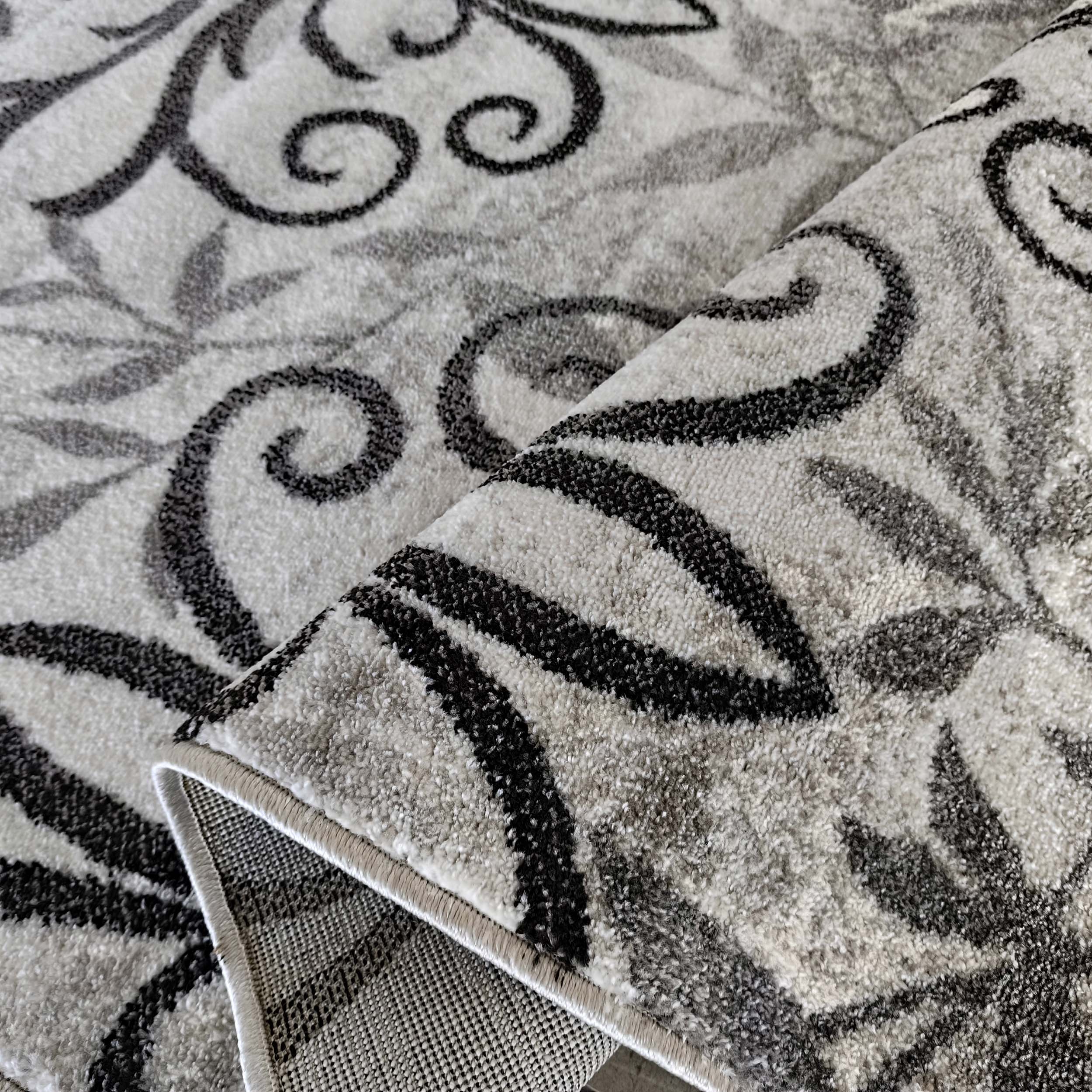 Tapis moelleux gris et blanc avec des motifs floraux abstraits noirs et gris foncés, dont une partie est roulée
