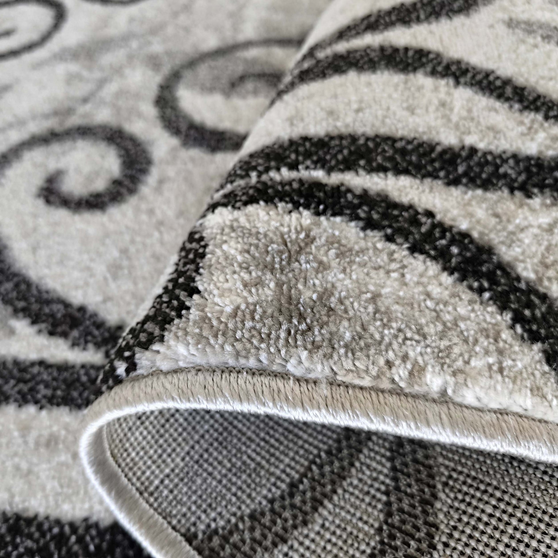 Détail d'un tapis moelleux gris et noir à motifs abstraits de spirales, partiellement roulé pour montrer son envers quadrillé