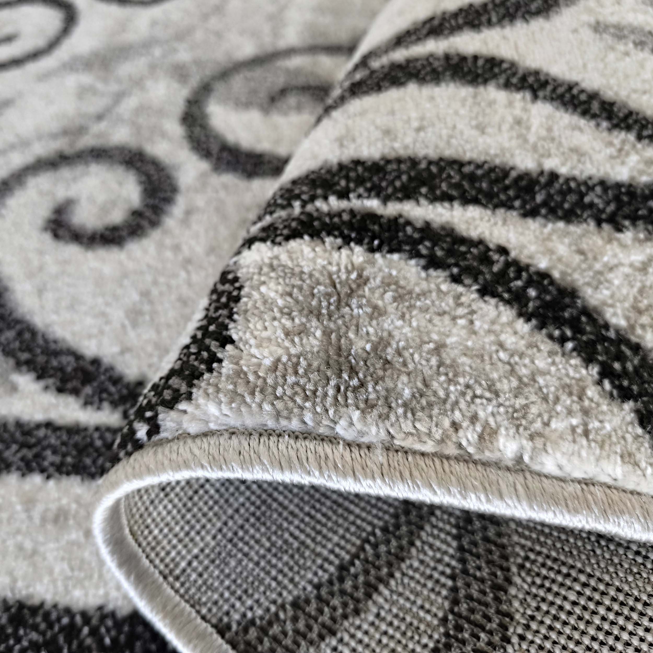 Détail d'un tapis moelleux gris et noir à motifs abstraits de spirales, partiellement roulé pour montrer son envers quadrillé