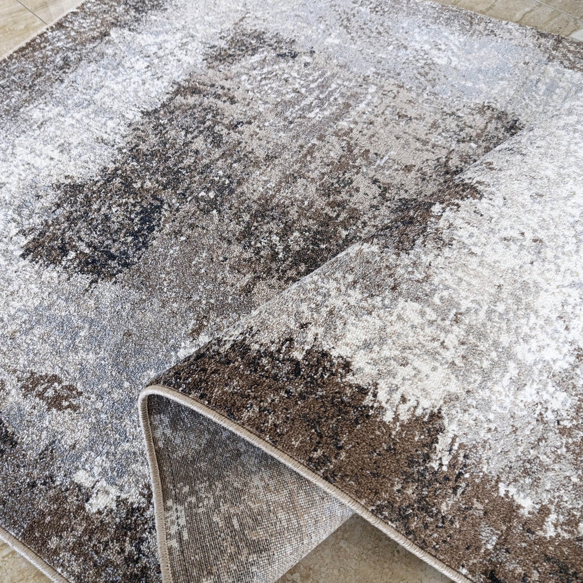 Tapis roulé avec un motif abstrait moucheté marron, blanc et gris, révélant sa face inférieure tissée beige et un sol carrelé