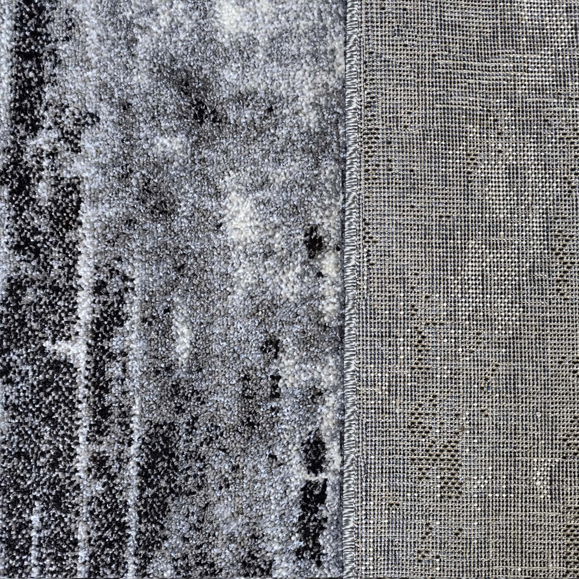 Gros plan d'un tapis à la surface moelleuse gris, blanc et noir aux motifs abstraits, et son envers en tissu gris clair tissé