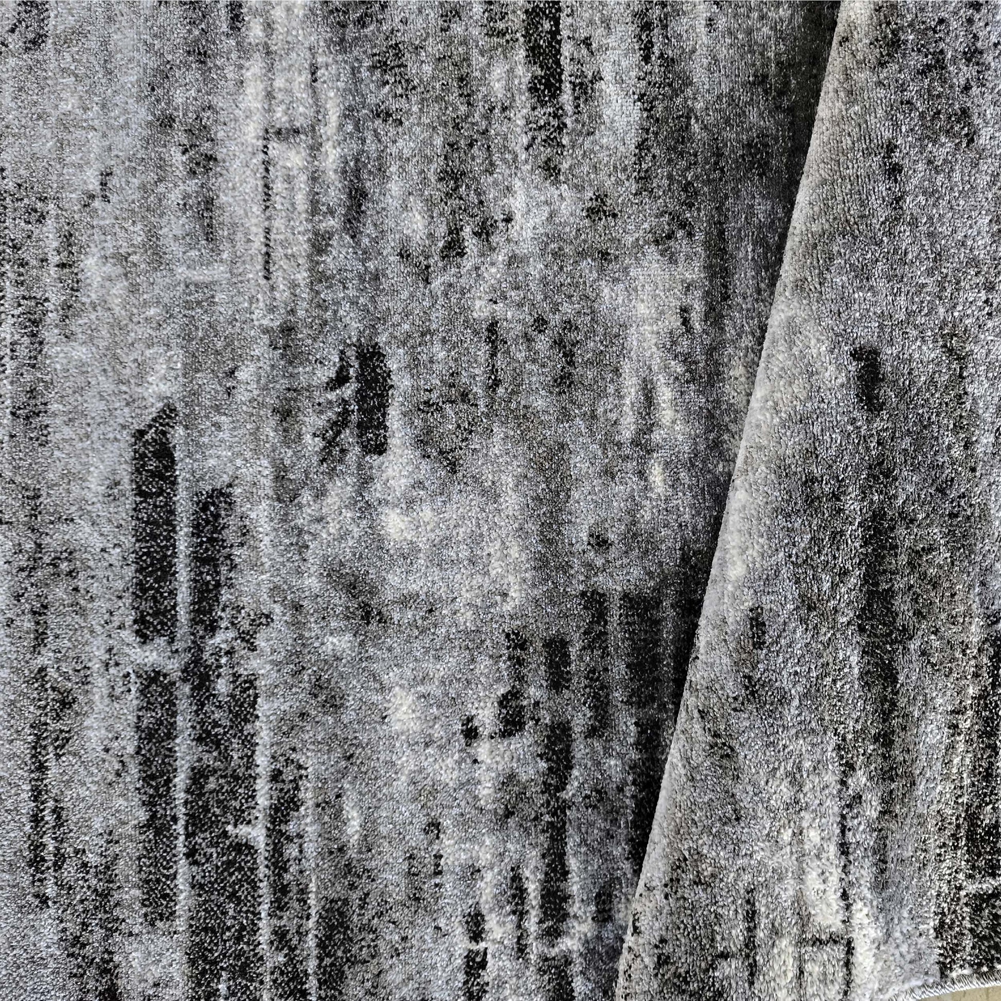 Tapis gris clair et foncé avec des zones noires, présentant une texture à poils courts et un motif abstrait usé, replié sur un côté