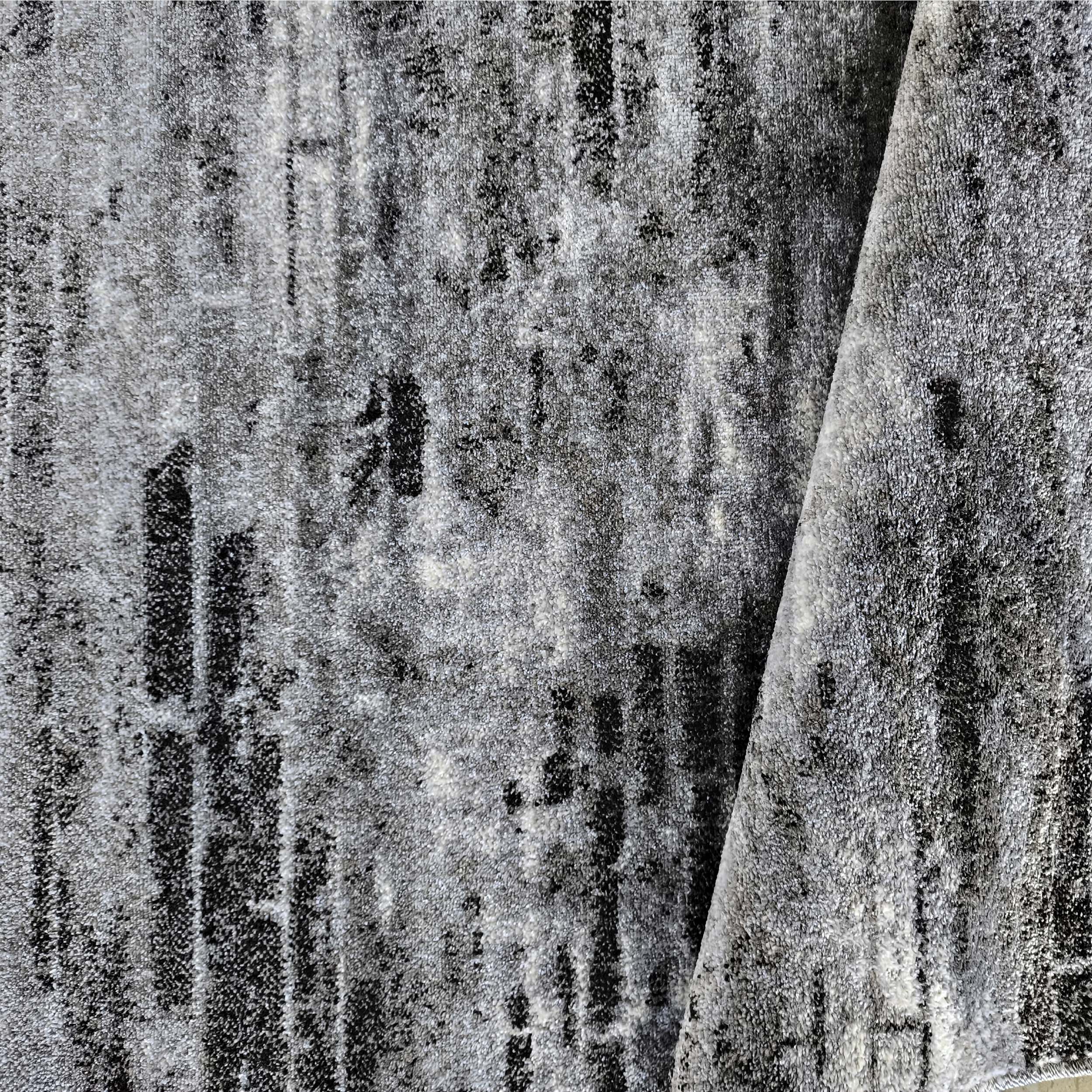 Tapis gris clair et foncé avec des zones noires, présentant une texture à poils courts et un motif abstrait usé, replié sur un côté