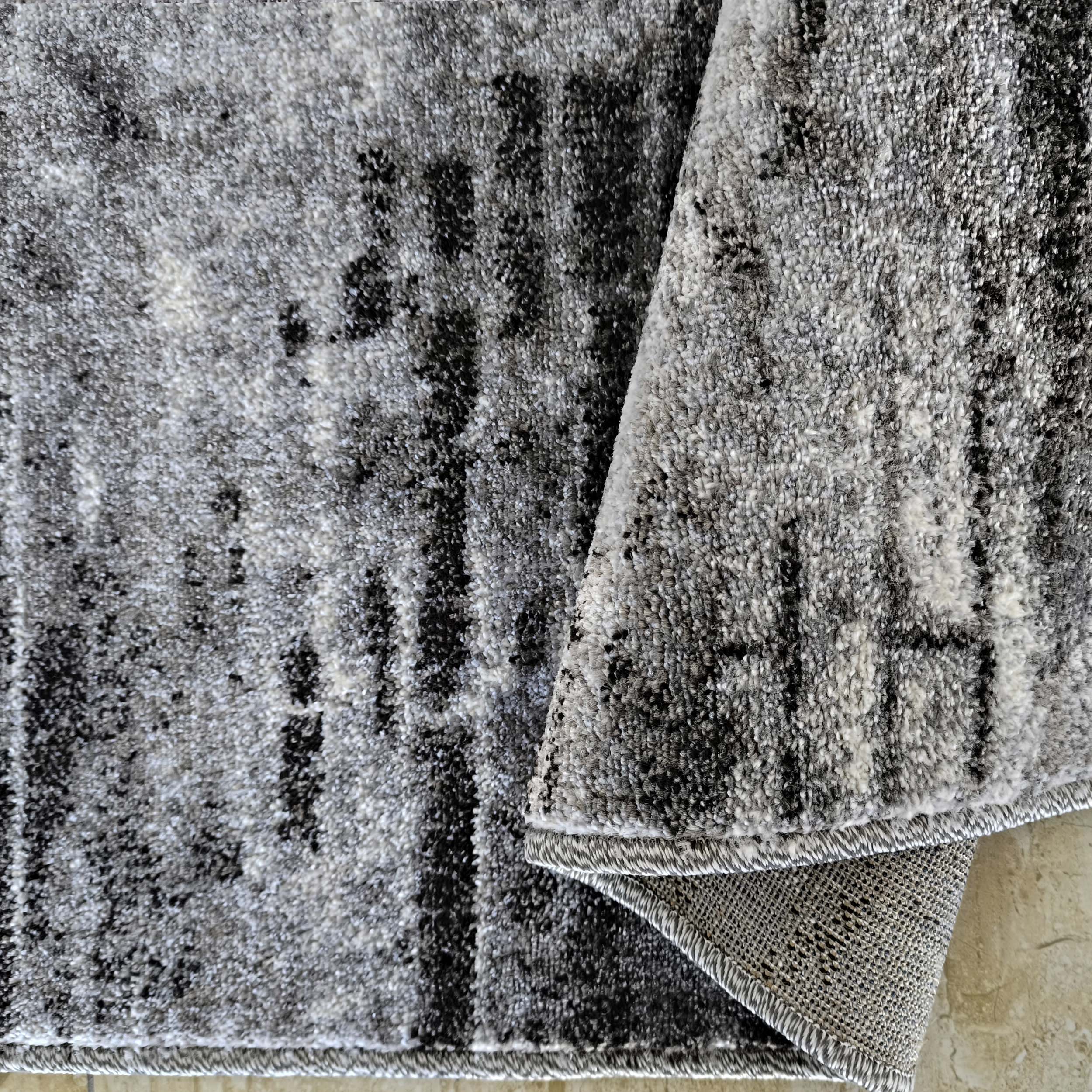 Tapis à poils longs aux motifs irréguliers gris, noir et blanc, partiellement roulé pour révéler l'envers tissé