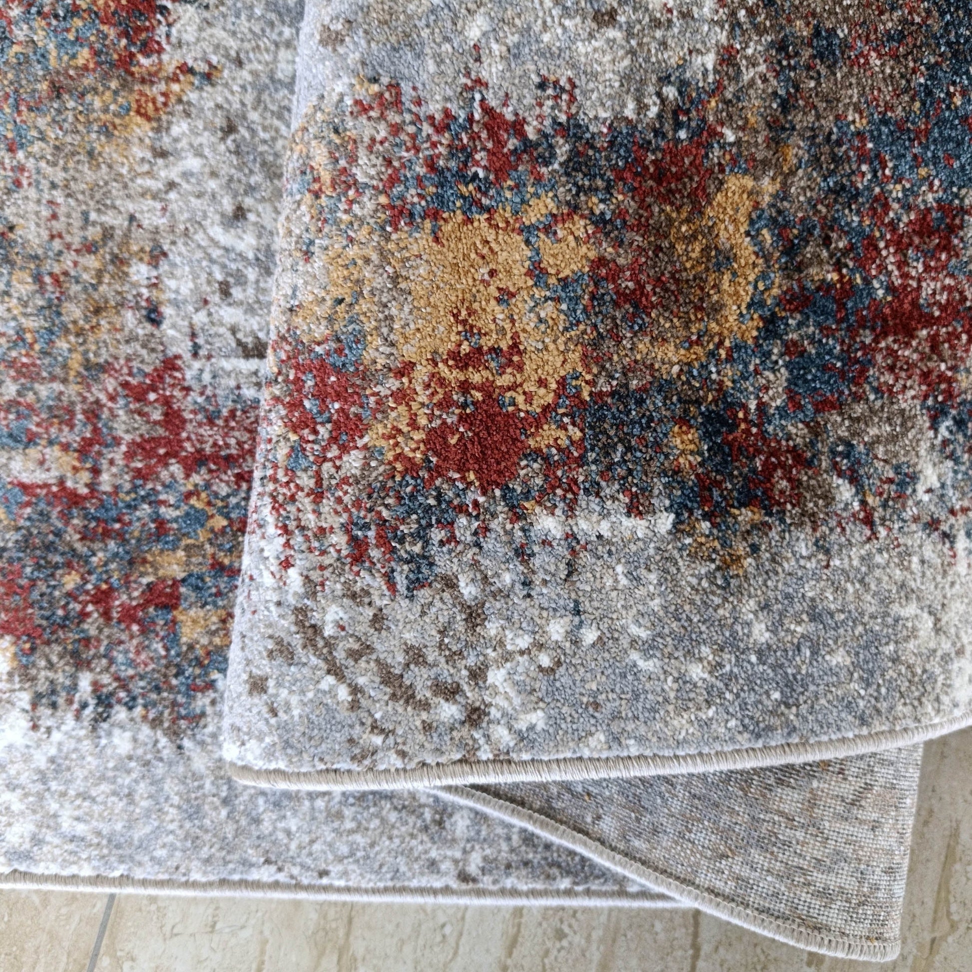 Tapis abstrait moucheté gris, blanc, marron, rouge, bleu, ocre, légèrement plié sur sol carrelé beige