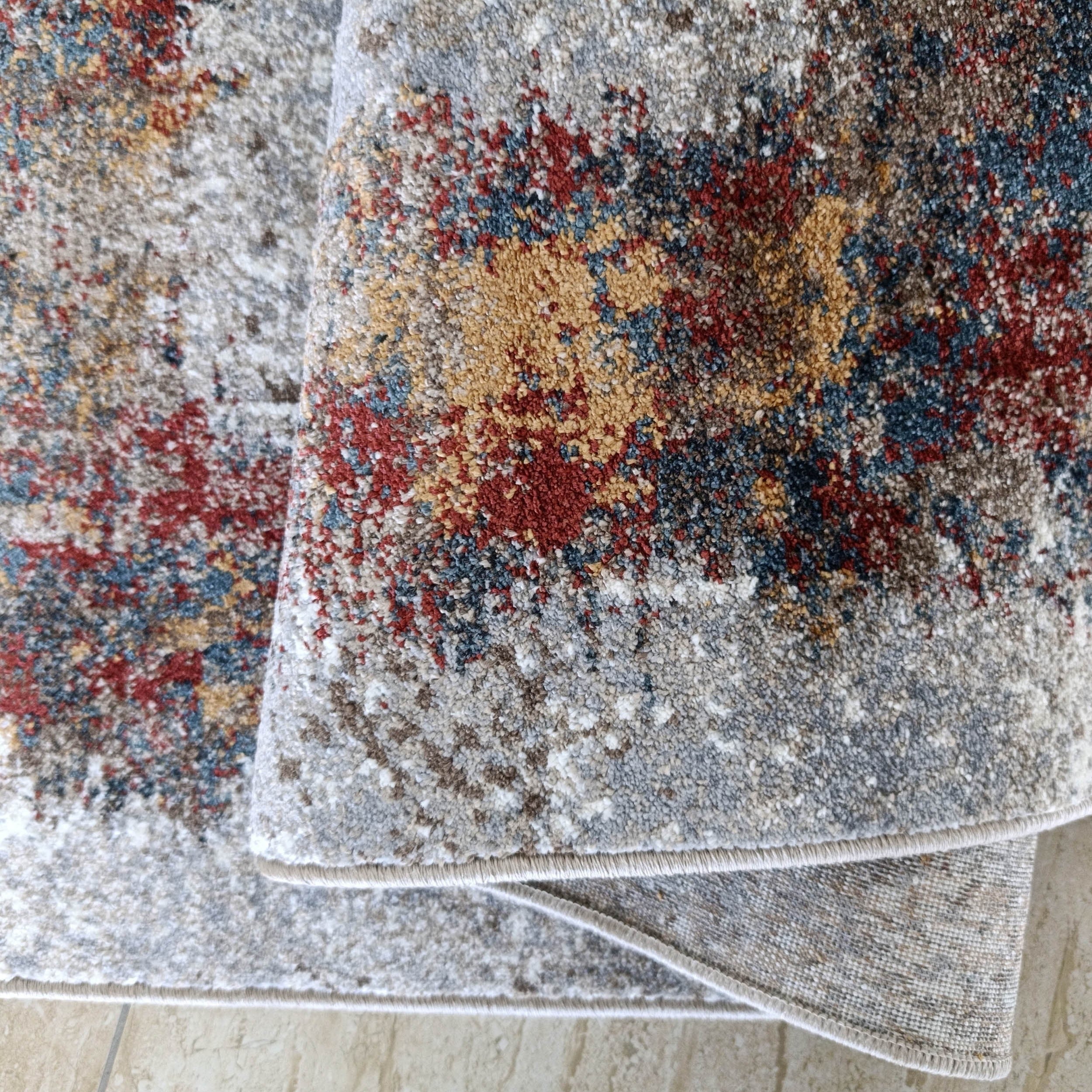 Tapis abstrait moucheté gris, blanc, marron, rouge, bleu, ocre, légèrement plié sur sol carrelé beige