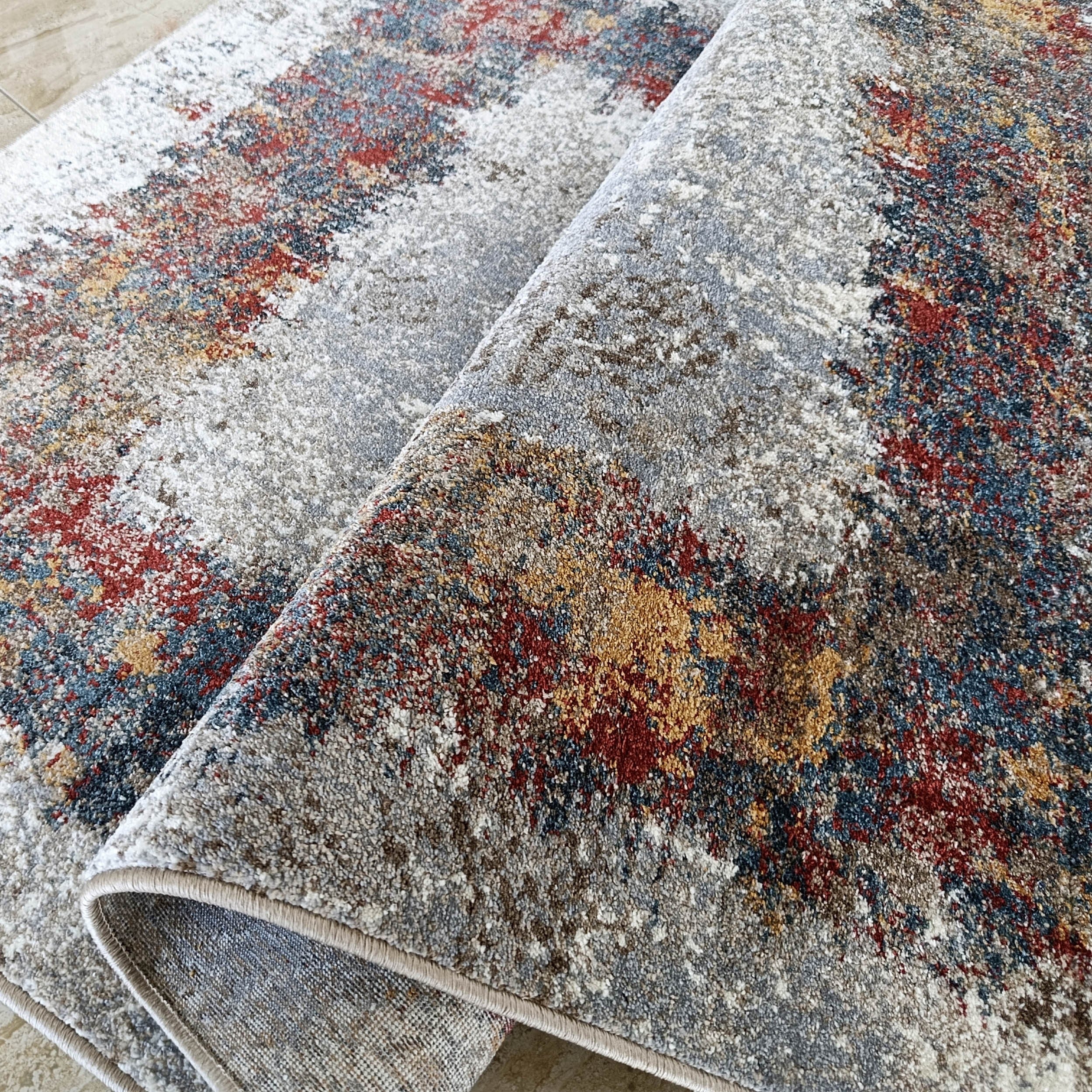 Tapis doux partiellement roulé aux motifs abstraits gris, rouge foncé, bleu marine et jaune moutarde sur un sol carrelé clair