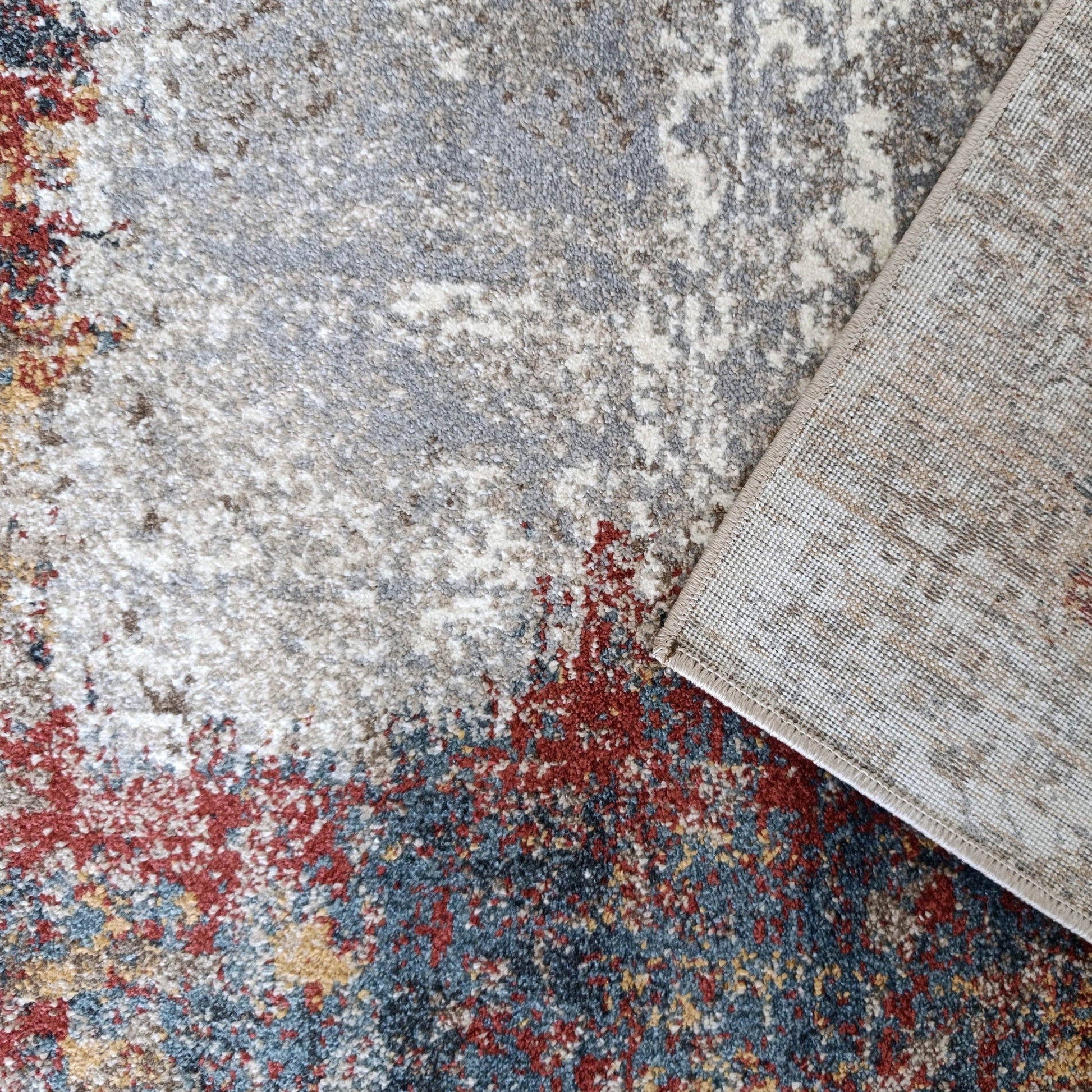 Tapis à motifs abstraits gris, blanc, rouge et bleu, partiellement retourné, dévoilant son envers tissé beige clair