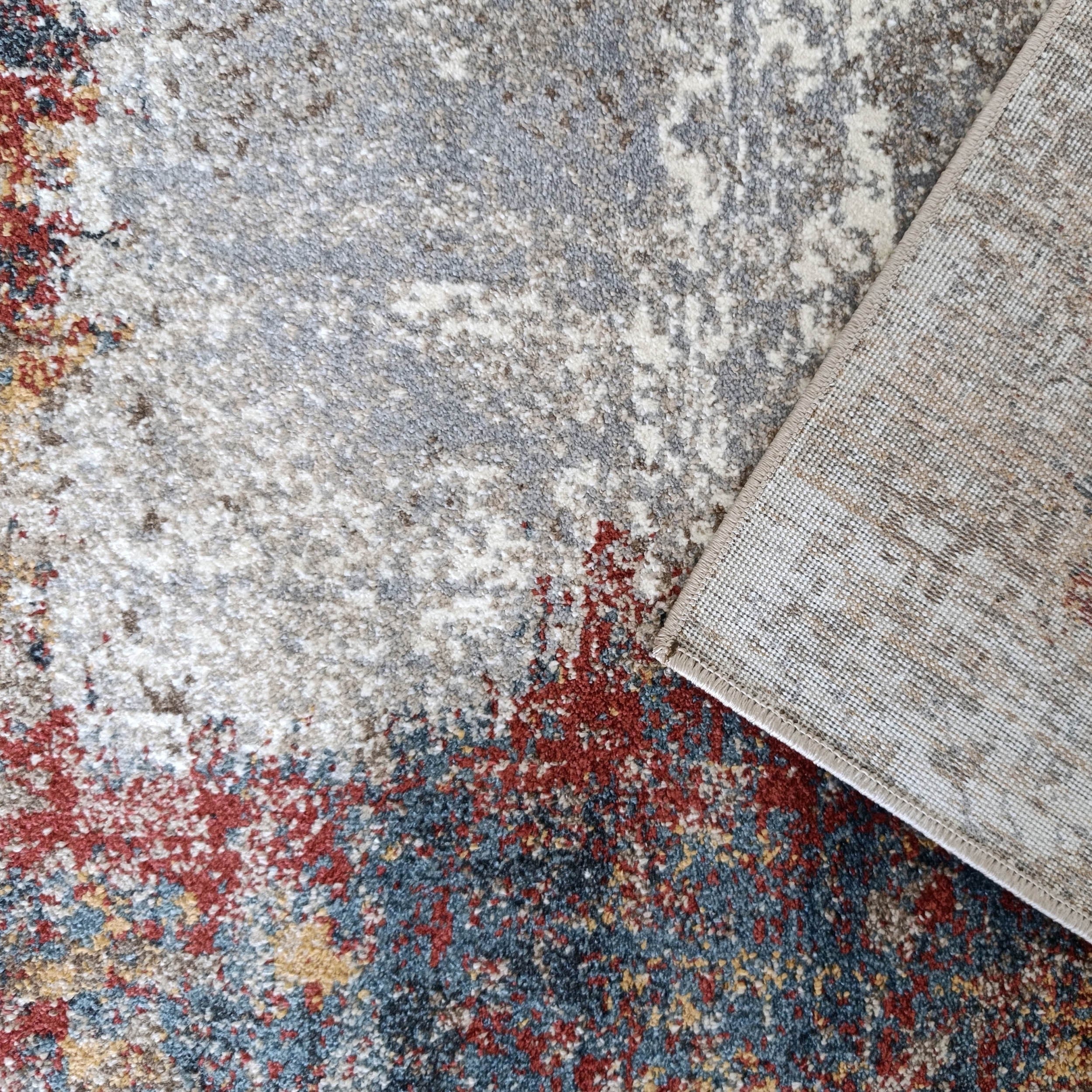 Tapis à motifs abstraits gris, blanc, rouge et bleu, partiellement retourné, dévoilant son envers tissé beige clair