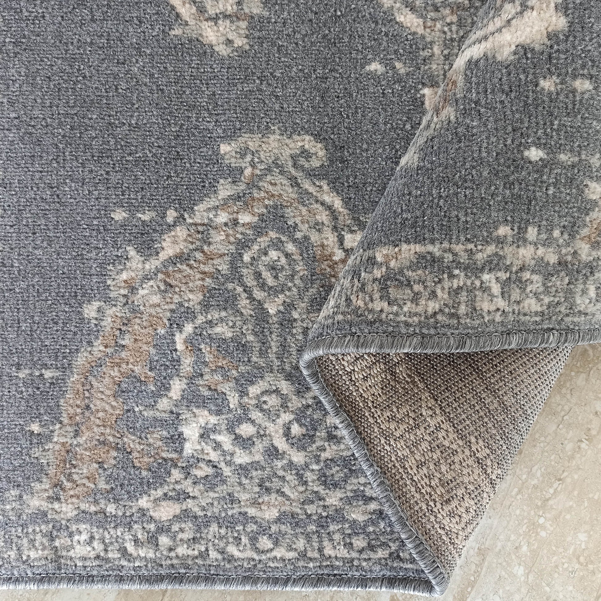 Tapis gris à motifs classiques beige, crème et marron clair, avec un bord surfilé gris et le dessous tissé exposé