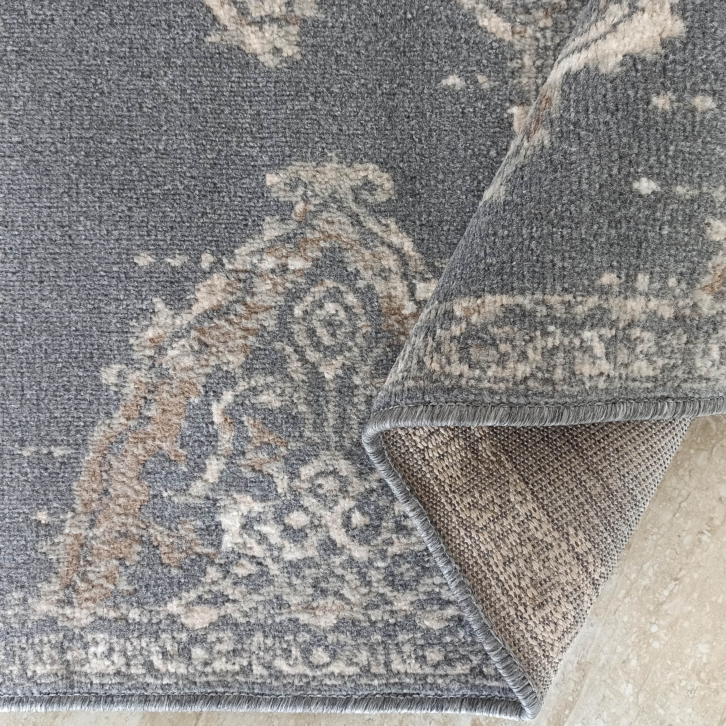 Tapis gris à motifs classiques beige, crème et marron clair, avec un bord surfilé gris et le dessous tissé exposé