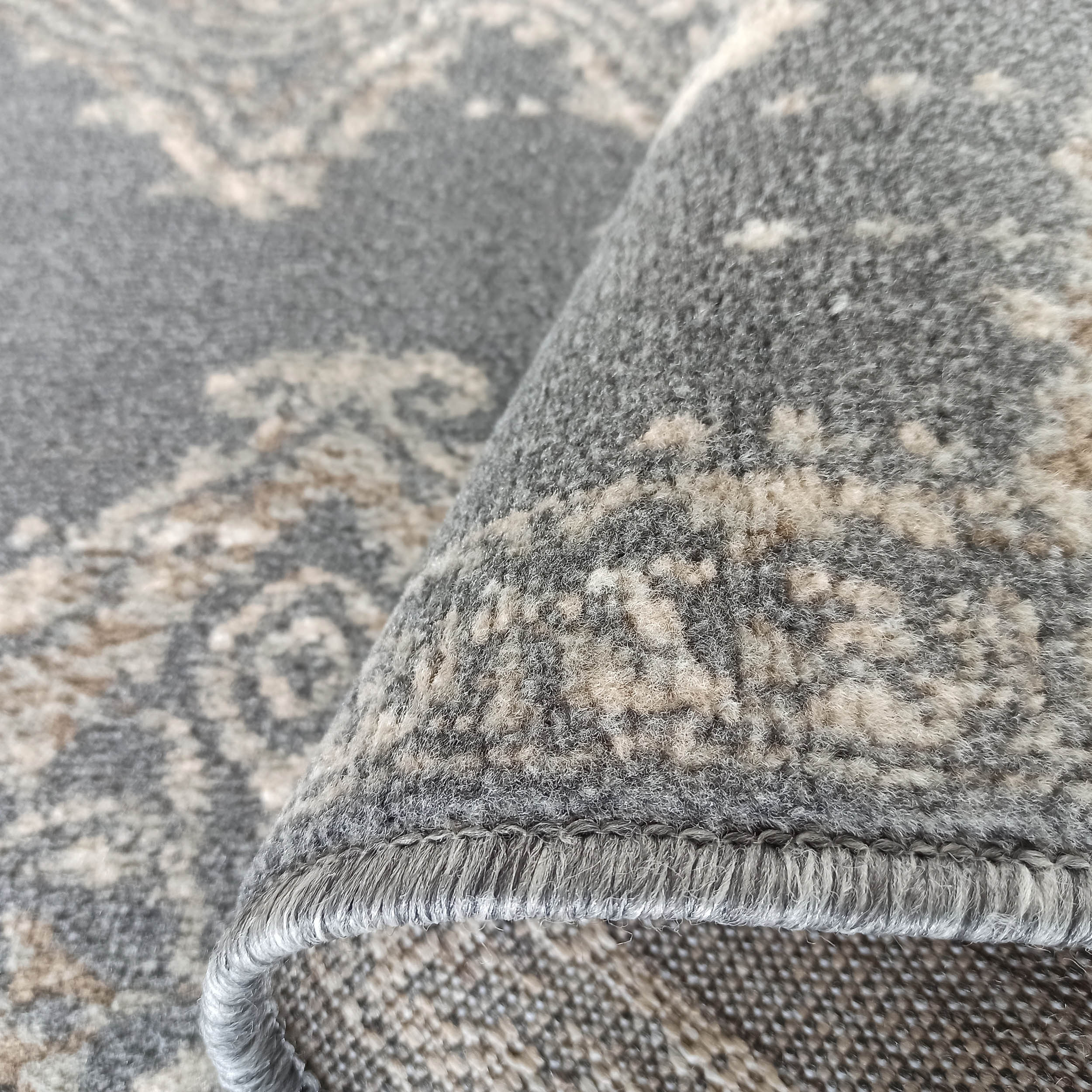 Tapis gris à motifs ornementaux beige clair, partiellement roulé, montrant sa texture douce et son bord cousu