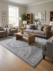 Tapis rectangulaire bleu-gris à motifs ornementaux crème et beiges, posé sur un parquet clair dans un salon lumineux