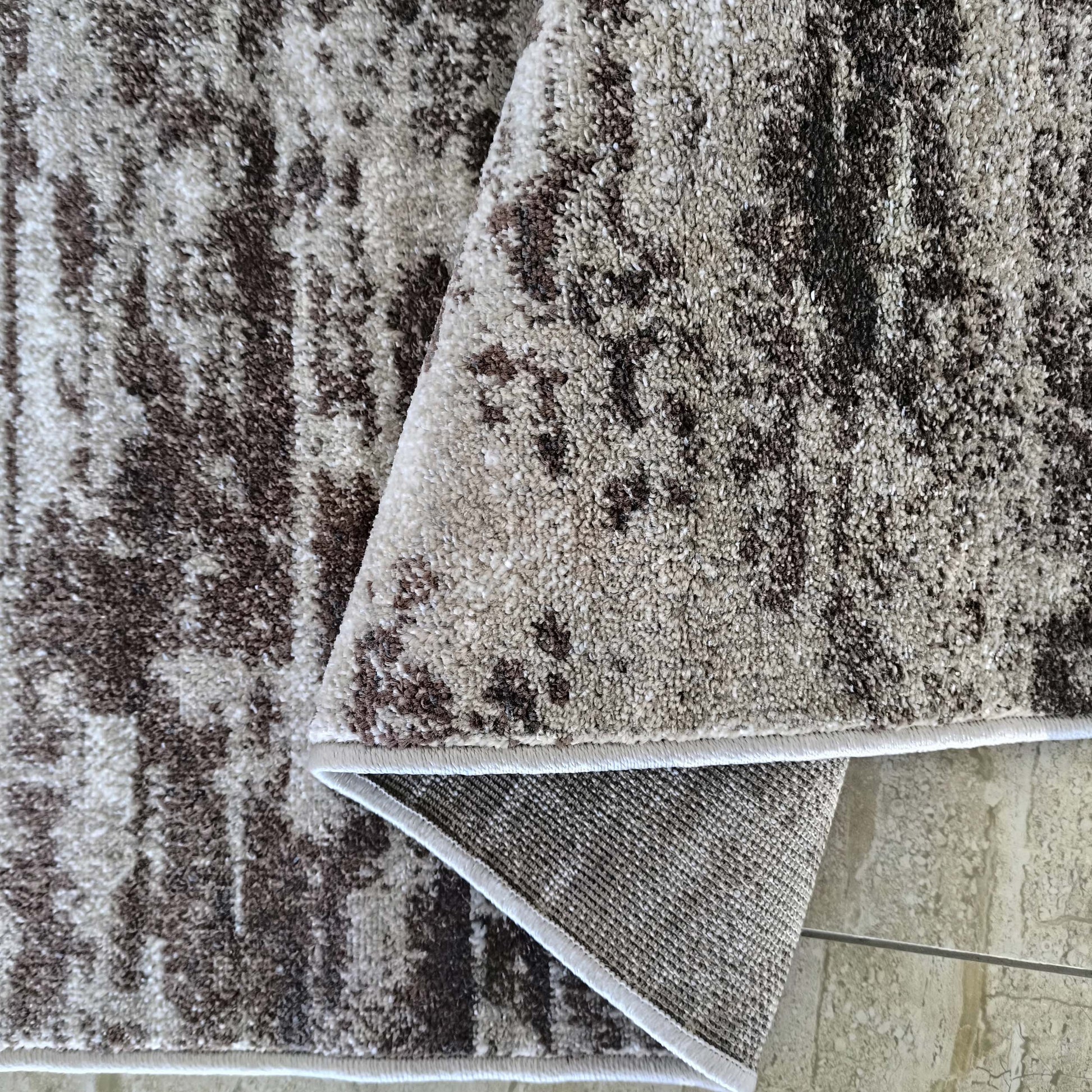 Tapis aux motifs abstraits tachetés marron foncé, beige et crème, à la texture moelleuse. Une partie est repliée, révélant son envers tissé
