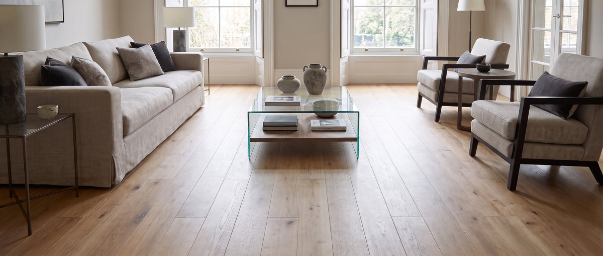 Spacieux salon moderne au sol nu, avec parquet clair, canapé beige, fauteuils gris et table basse en verre